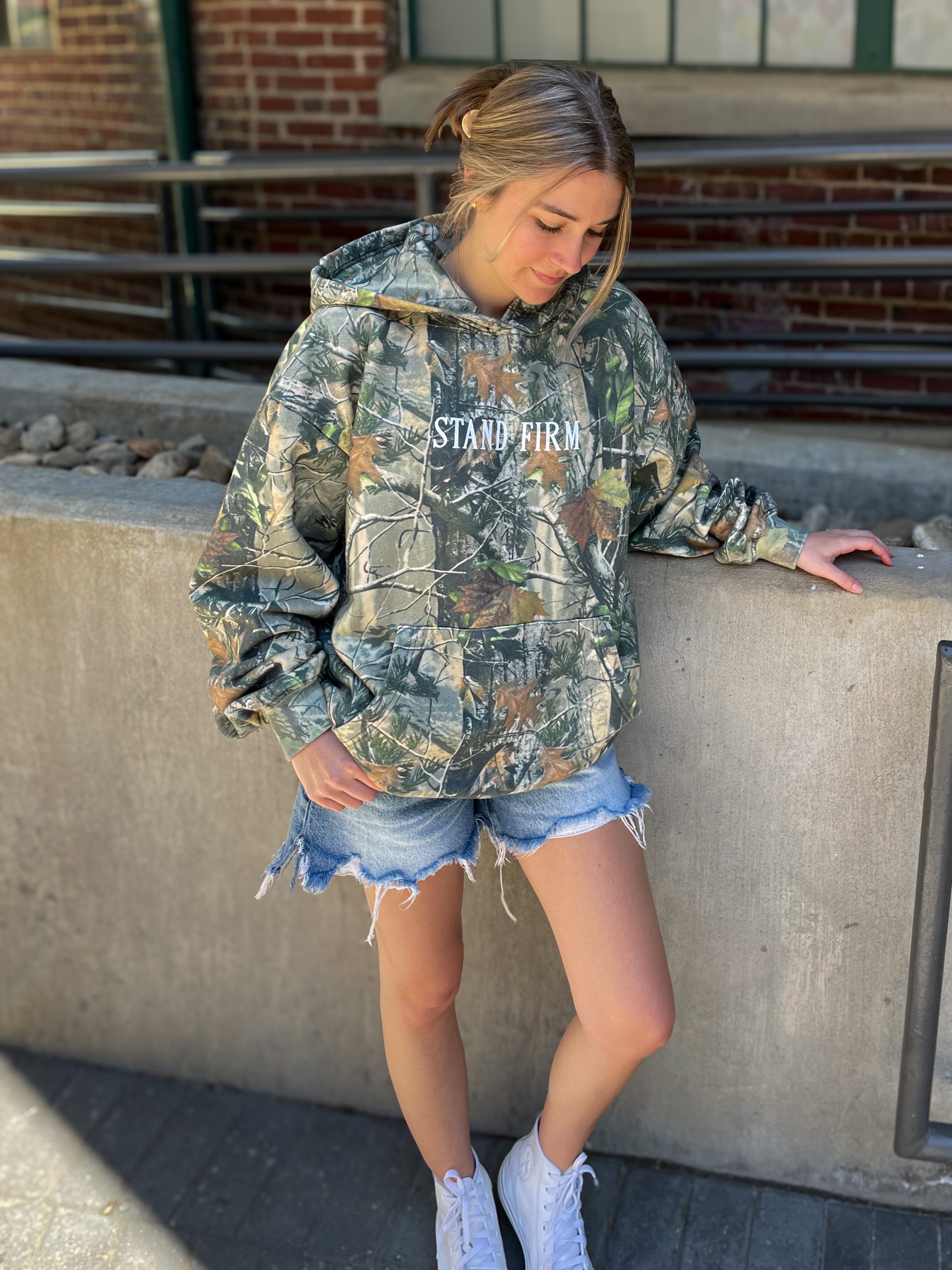 a woman leaning against a wall wearing a camouflage hoodie
